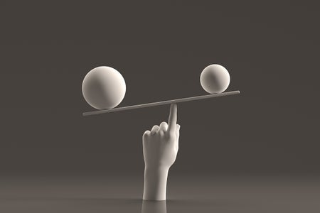 Still life of a white hand sculpture balancing different sized white spheres on a white plank shown against a grey backdrop.