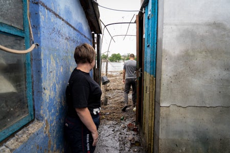 Two people stand between concrete buildings, facing a flooded garden