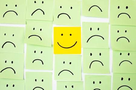 A wall covered in green sticky notes with sad faces drawn onto them and one yellow stick note with a happy face drawing