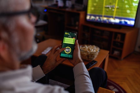 Mature person sitting on a sofa at home, watching soccer game while using a smart phone for sports betting