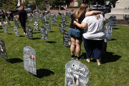 Two mourners embrace in a city park standing amongst a representative graveyard installed as a temporary memorial for individuals in the community who died by overdose