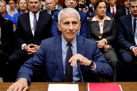 Dr. Anthony Fauci in blue suit and microphone at hearing with people shown in background.