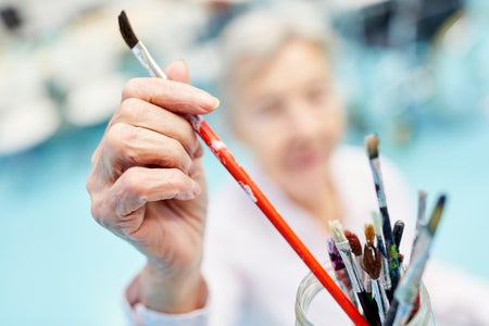 Close up of an older person's hand removing a painting brush from a glass jar holding multiple other brushes