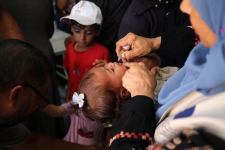 A small child receives an orally administered polio vaccine at the center of the image, surrounded by other adults, children, and medical workers in a field hospital in Gaza